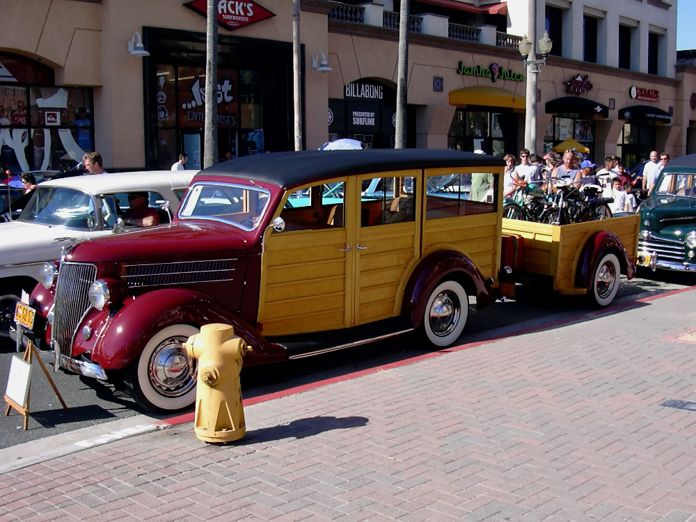 Auto-generated description: A vintage car with a wooden exterior is parked on a street next to a trailer, surrounded by people and other classic cars.