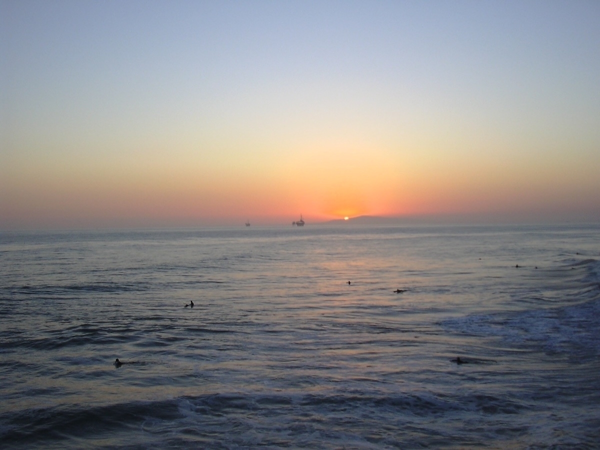 Auto-generated description: The photo captures a serene ocean scene at twilight, with surfers in the water and the sun setting on the horizon.