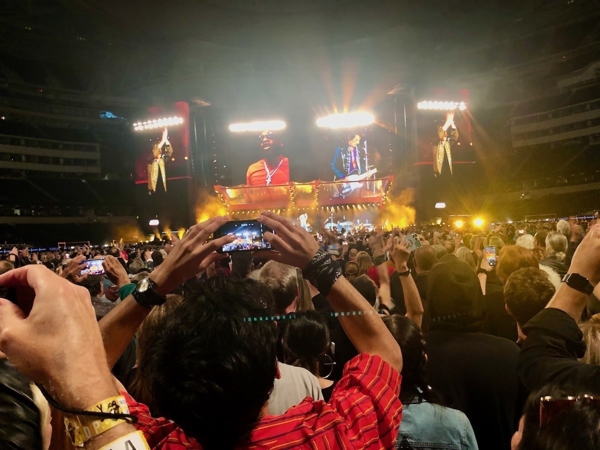 Auto-generated description: A large crowd at a concert captures the moment with their phones as performers play on a brightly lit stage.