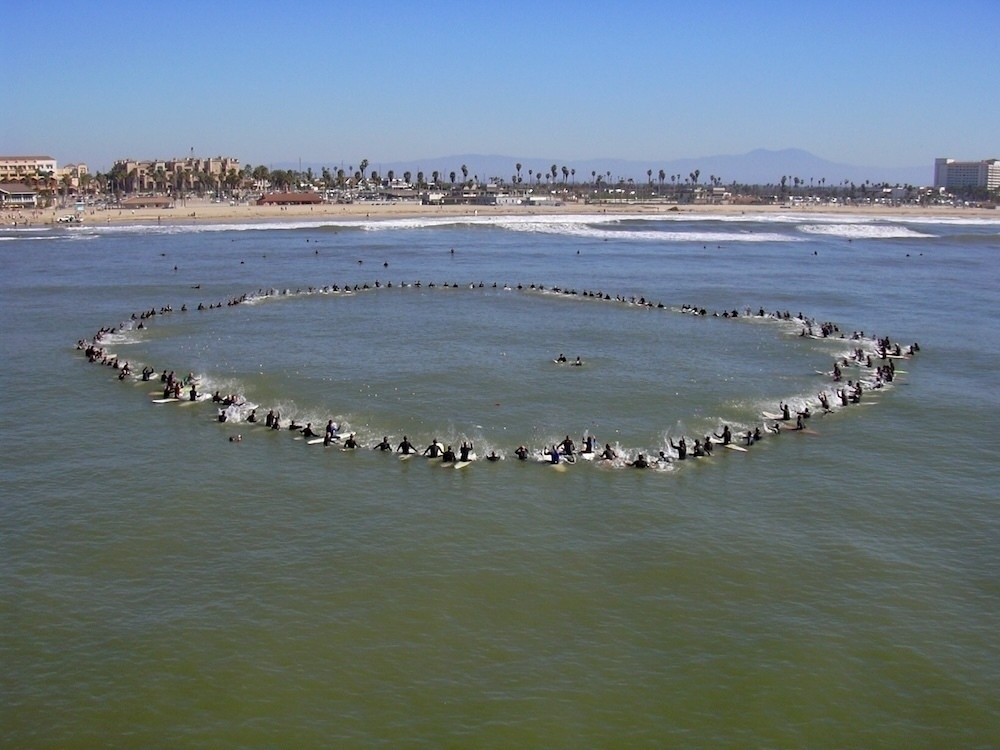 Auto-generated description: A large group of surfers forms a circle on the water near a sandy beach.
