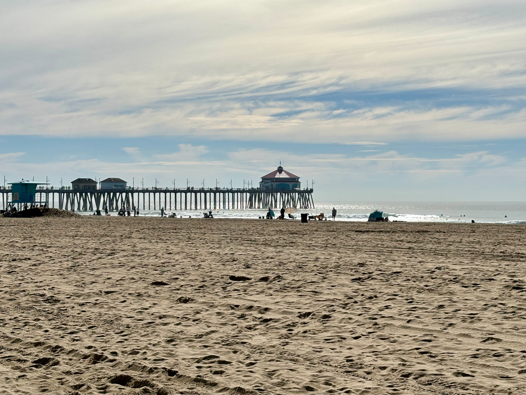 A sandy beach stretches out towards a distant pier extending over the ocean, with a few people relaxing near the water under a partly cloudy sky.
