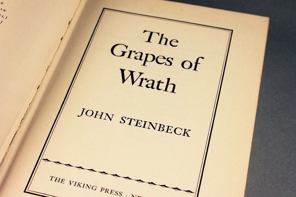 An open book displays the title page of 'The Grapes of Wrath' by John Steinbeck, published by The Viking Press.