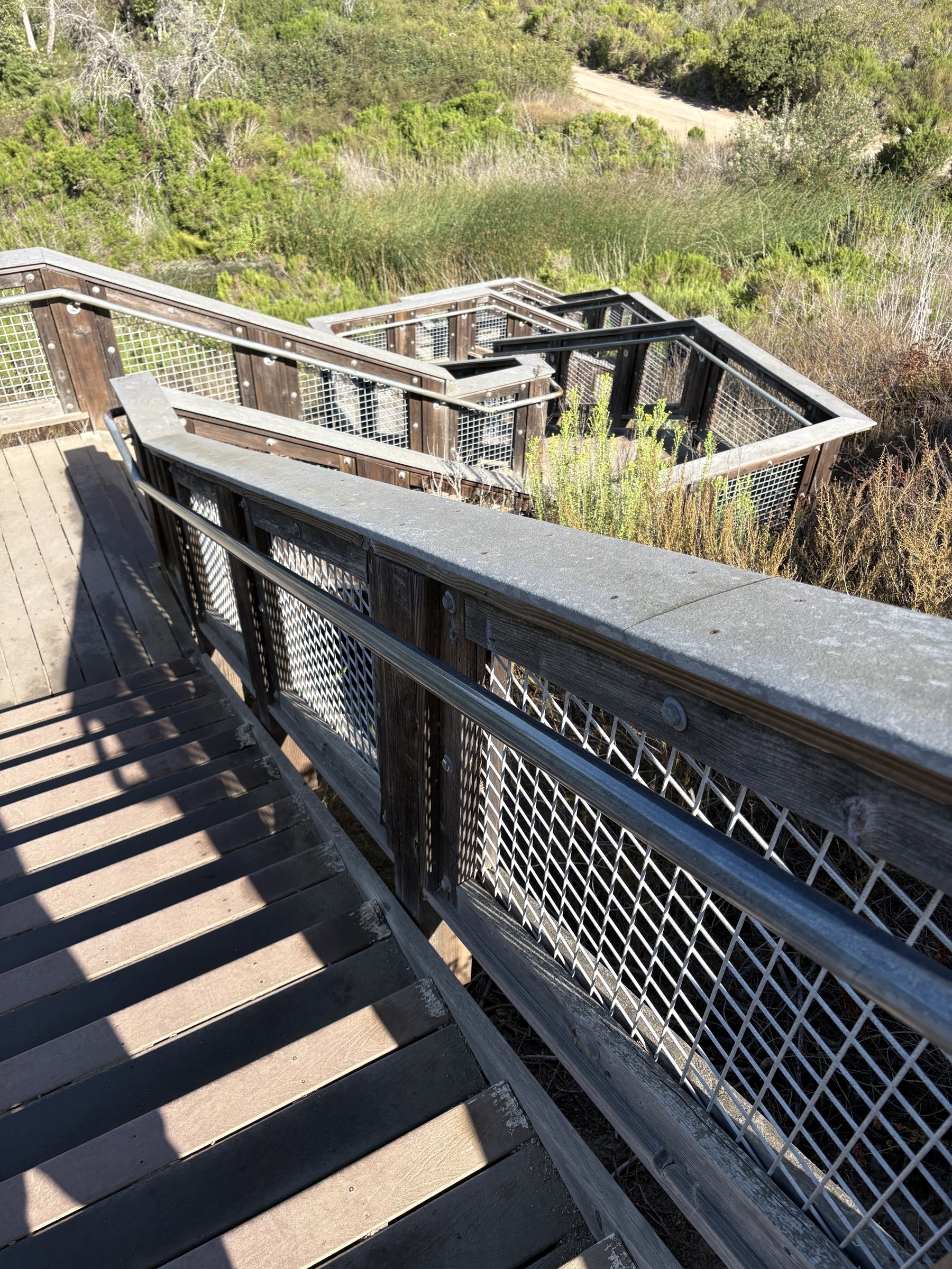 A wooden staircase with metal railings leads down through a natural, green landscape.